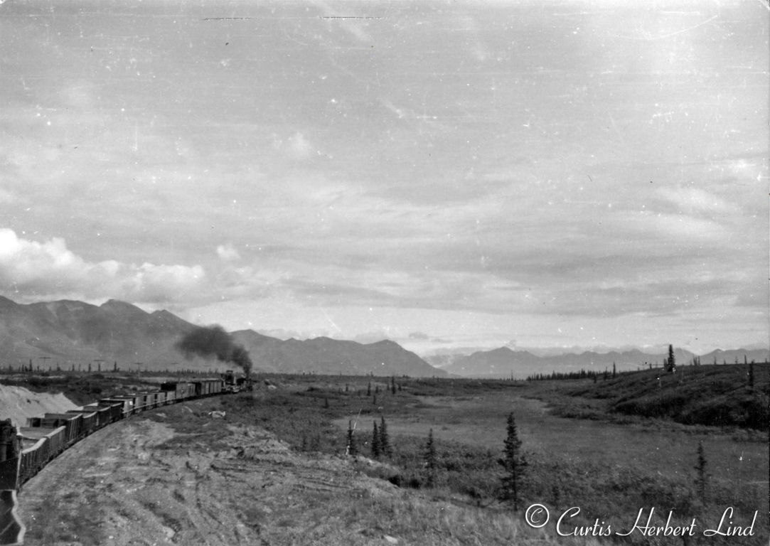 Somewhere in the Broad Pass area this MOW train is headed up by a steam engine. Right behind the loco is a large track mounted shovel on a flat car. It appears to be moving to a new work site with a string of well used Hart Convertible Gondolas and a menagerie of odd gone. The car in the foreground has seen better days as a result of that shovel banging away on it. This is after 1948 because they are already using a Pullman Troop sleeper as a camp car. Notice the telecom lines along the ridge line. In those days there were no roads in this area. Is that white line going across the swamp a pipeline just laid on the ground? 
